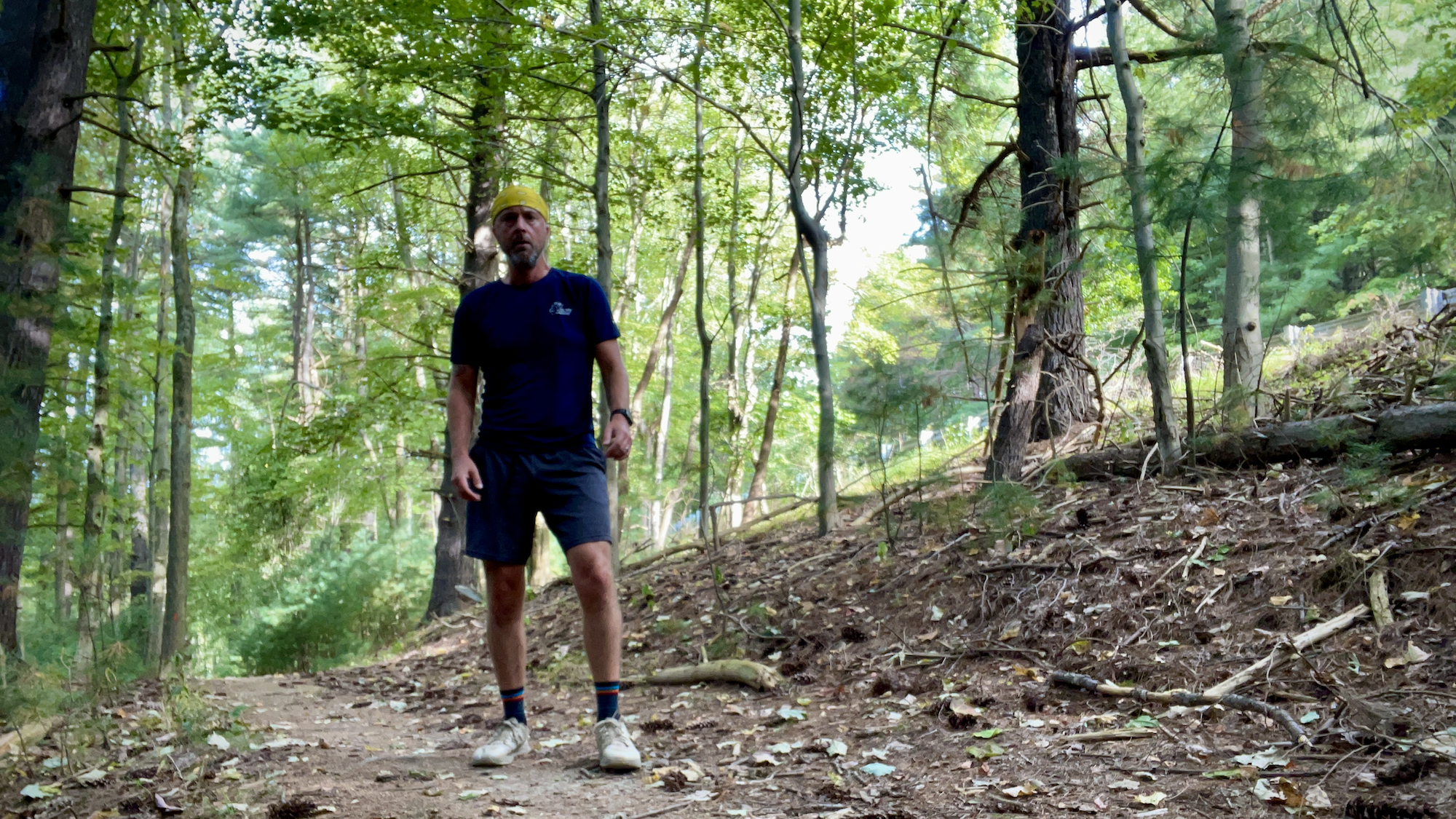 A man in a blue shirt, grey shorts and yellow bandana standing on a trail.