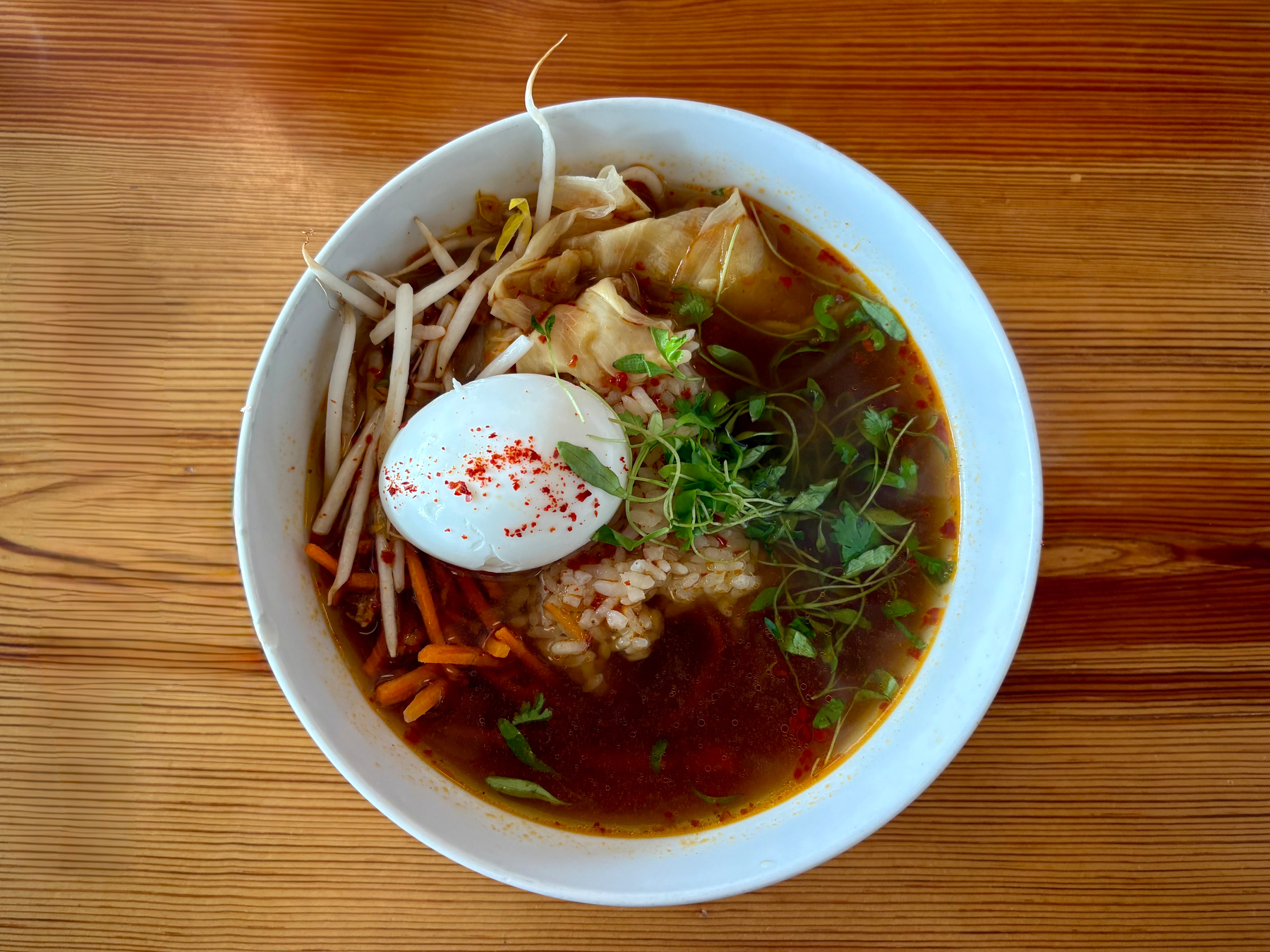 A bowl of soup contains a boiled egg, bean sprouts, leafy greens, rice, and a flavorful broth with red seasoning sprinkled on top.