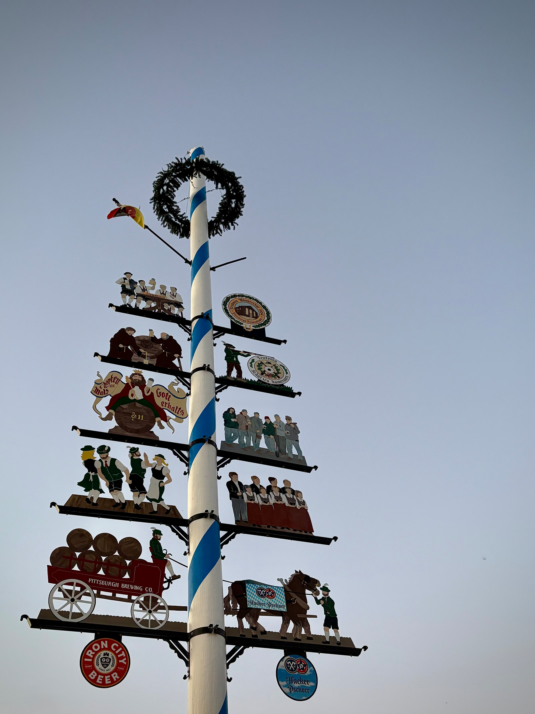 A tall maypole decorated with traditional figures, symbols, and signs, including people dancing, a horse-drawn barrel cart, and brewery logos, stands against a clear sky.