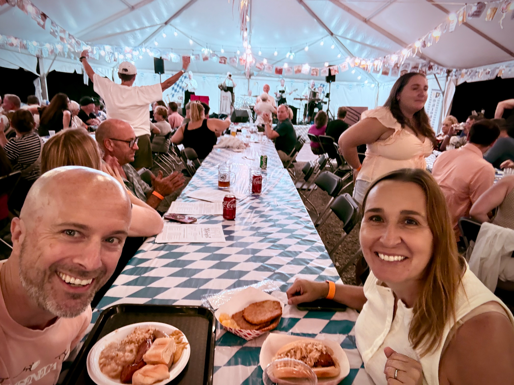 A lively gathering under a decorated tent features people enjoying food and drinks at long tables, with a smiling couple in the foreground.