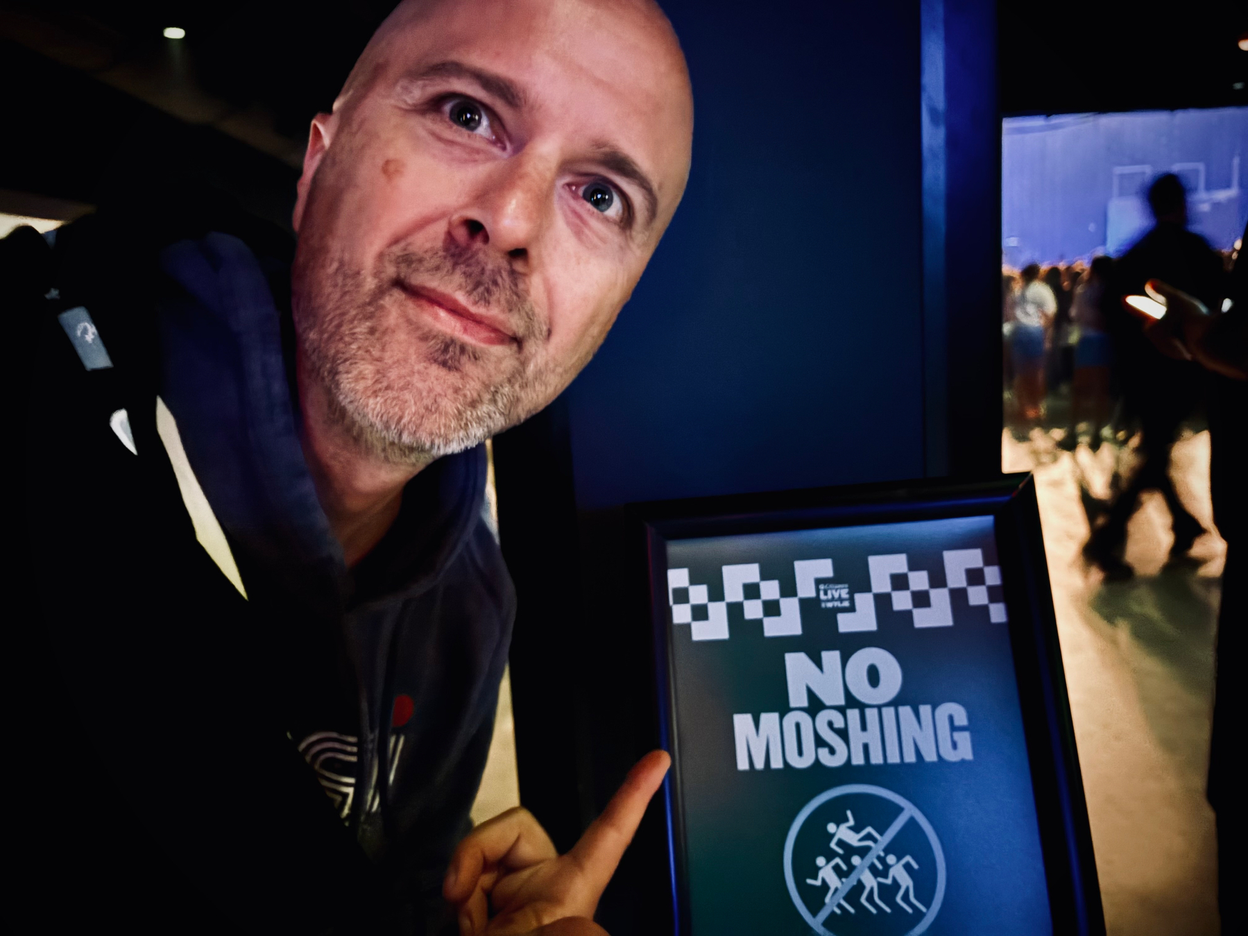 A man is pointing at a sign that says NO MOSHING inside a dimly lit venue with people in the background.